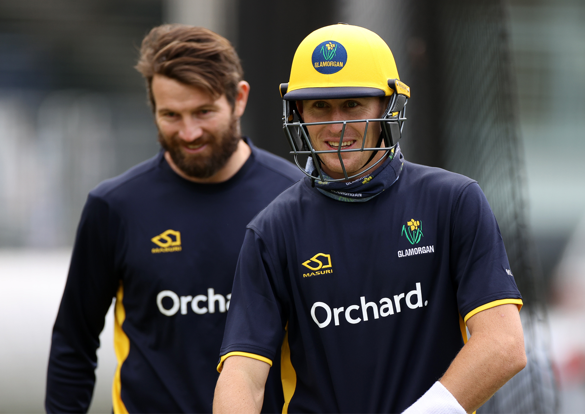 Two smiling men in Glamorgan County Cricket Club kit. One man is wearing a yellow cricket helmet