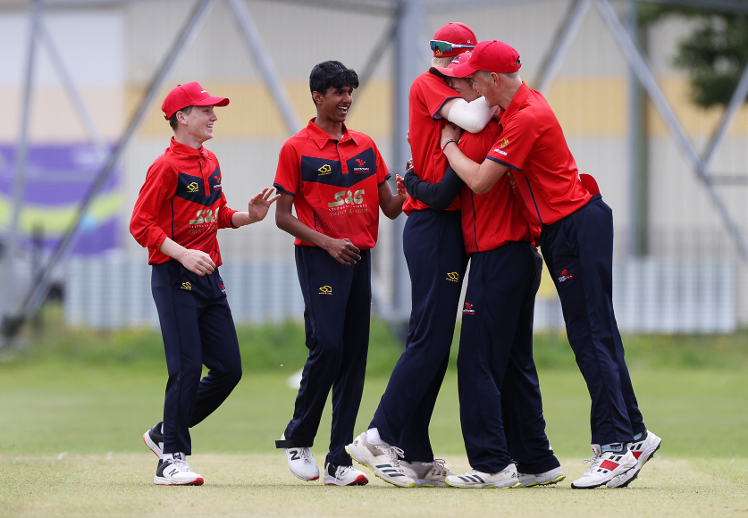 5 players in Cricket Wales kit smiling and embracing on a green