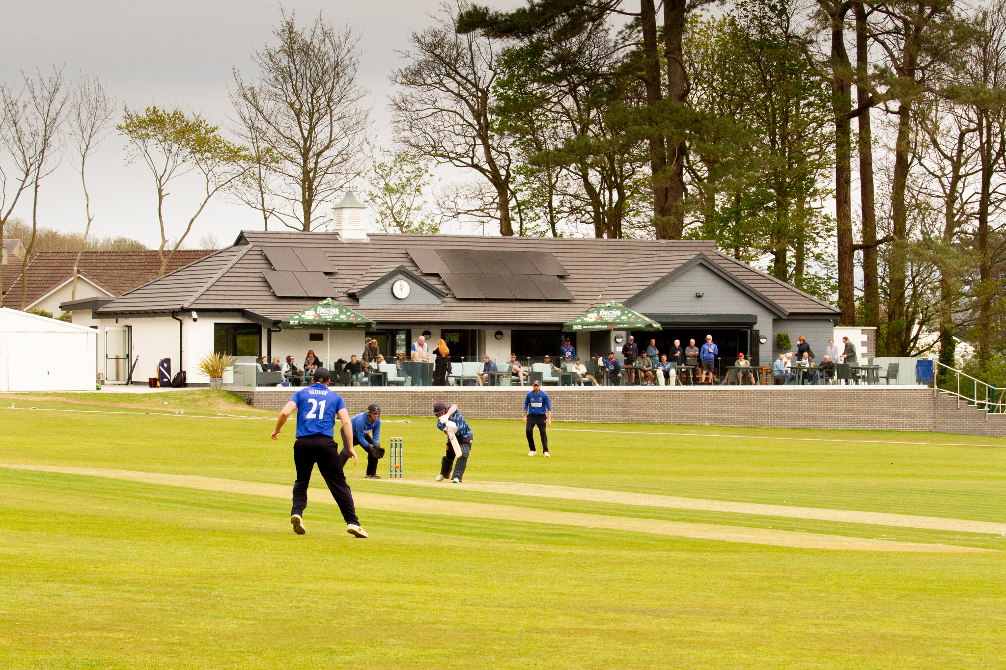 Cricket players in blue kit playing in front of a white club house