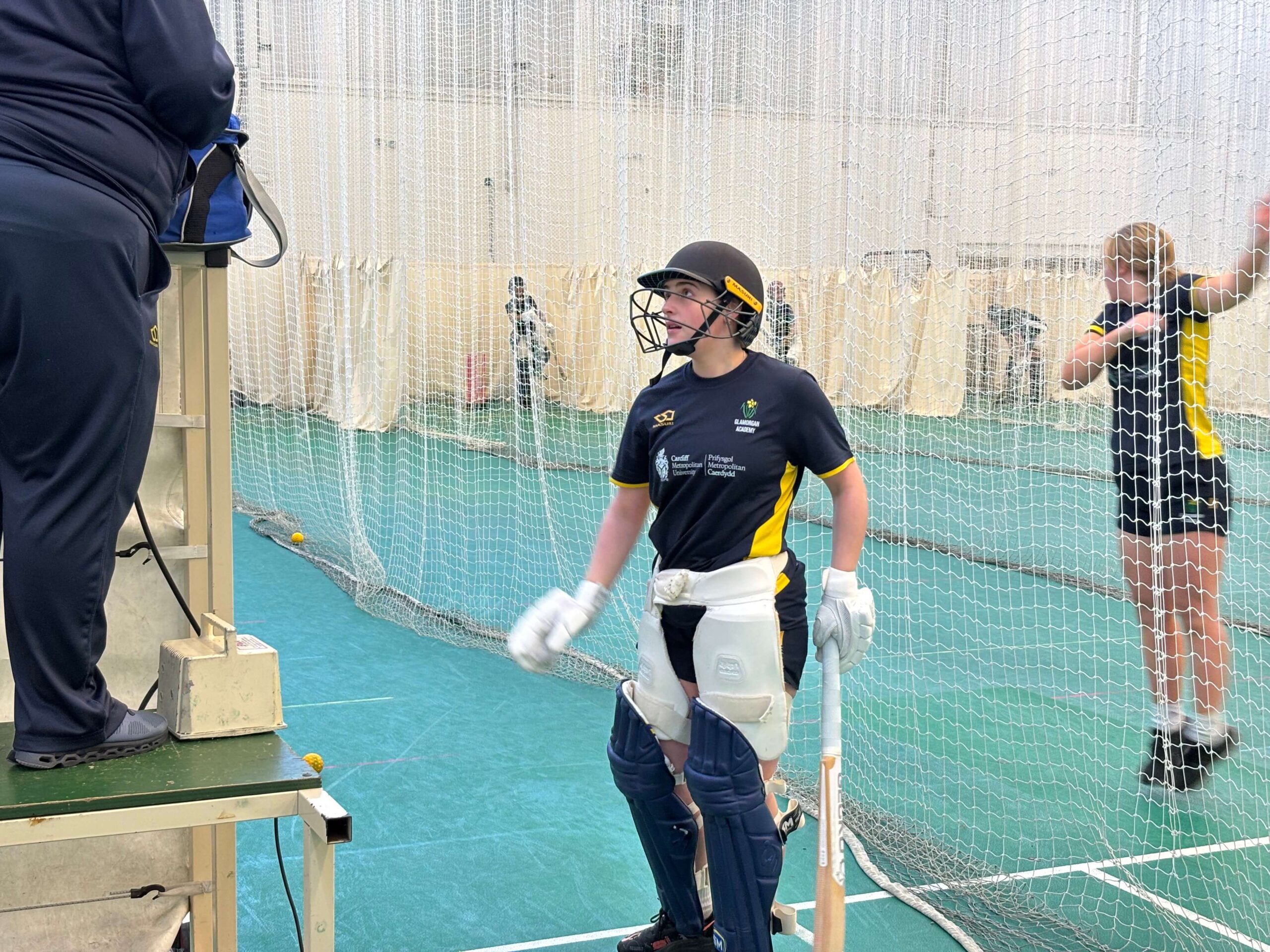 Young women in Glamorgan County Cricket Club kit holding bat, looking up in practice court